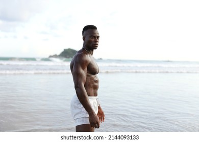 Half Body Shot Of A Handsome Young Man Standing On A Ocean Beach.shirtless Wearing Boxer Shorts, Showing Muscular Fit Body.Looking For Camera