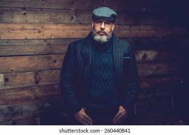 Half Body Shot Of An Old Goatee Man In An Elegant Dark Formal Wear In Front A Wooden Wall With Hands On The Pocket While Facing At The Camera Seriously.