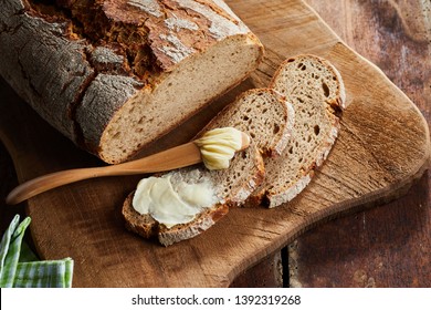 Half buttered slice of rye bread with spreader or butter knife on a wooden cutting board with crusty loaf viewed top down in close up