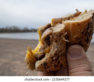 Half Eaten Breakfast Bagel Sandwich Held Up With Male Hand On A Cloudy Day By The Shoreline.