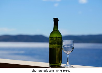 Half Empty Bottle And One Wineglass With White Wine Standing On Railing Of Balcony On Blurred Seascape And Blue Sky Background.