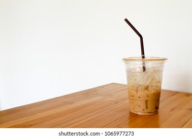 A Half Empty Cup Of Iced Caramel Latte Coffee In A Transparent Plastic Cup With Plastic Drinking Straw On Wooden Table And White Wall Background.