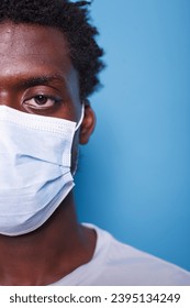 Half face, close-up of black man with mask staring at camera, against blue background. Cropped image of african american individual wearing face mask for protection from coronavirus. - Shutterstock ID 2395134249