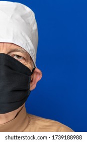 Half Face Senior Adult Doctor Dressed Black Surgical Face Mask, White Medical Cap, Standing Against Blue Background And Piercingly Looking At Camera. Close-up Head Shot. Concept Preventive Protection.