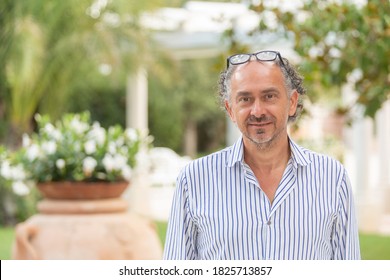 Half Figure Portrait Of Tanned Middle Aged Italian Man Wearing Eyeglasses On His Head With Jeans And Blue Striped White Shirt Near A Terracotta Amphora In A Green Park With Grass Flowers And Trees