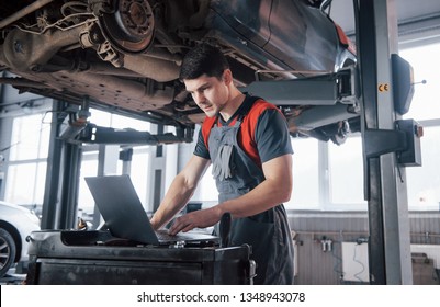 Half of the job is done. Man at the workshop in uniform using laptop for his job for fixing broken car. - Shutterstock ID 1348943078