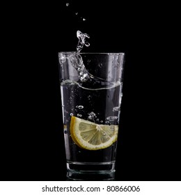 Half Of Lemon Falling Down In Glass With Water On Deep Black.  Beautiful Glass With Sparkling Water Or Other Transparent Drink And A Slice Of Lemon On Black Background