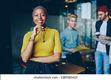 Half length portrait of african american female leader of working team standing on frontage while her colleagues talking on background prosperous business woman in casual wear  looking at camera