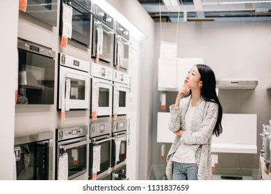 Half Length Portrait Of Asian Woman Choosing New Oven In Furniture Store