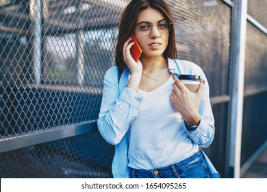 Half length portrait of attractive brunette woman in trendy apparel making mobile phone talk in roaming standing on urban setting,serious hipster girl using smartphone for calling holding coffee to go