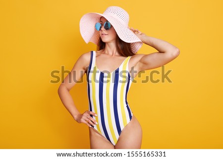 Half length portrait of beautiful slim Caucasian woman with sporty body, wearing bright striped swimsuit and beach hat posing with her hand raised, looking thounghtfully aside, isolated on yellow wall