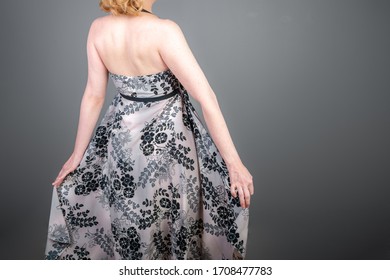 Half Length Portrait From Behind Of Woman Wearing Cute Dress. Long, Flowing Print Dress With Empire Waist. Woman Is Spinning.Studio Shot With Gray Background.