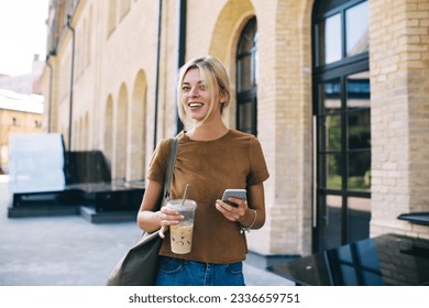 Half length portrait of Caucasian blogger with digital mobile phone and ice coffee smiling at camera during weekend time in city, happy millennial hipster girl with modern cellphone posing outdoors - Powered by Shutterstock