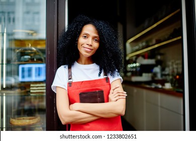 Half length portrait of cheerful african american professional barista in red apron smiling at camera.Positive dark skinned female waitress with crossed hands standing at entrance to own coffee shop