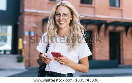 Half length portrait of cheerful caucasian female in trendy wear spending time on street using smartphone, beautiful millennial hipster girl blogger looking at camera in town holding mobile phone