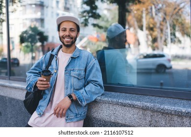 Half Length Portrait Of Cheerful Hipster Guy Dressed In Trendy Denim Clothes Smiling At Camera During Coffee Break In Touristic City, Happy Traveller In Cap Holding Takeaway Caffeine Beverage