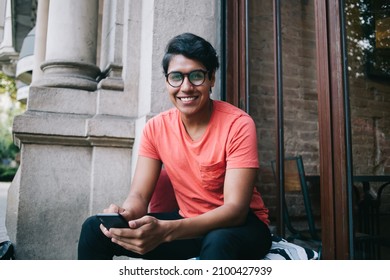 Half length portrait of cheerful male blogger with cellular gadget in hands resting at sidewalk area and smiling at camera during leisure pastime in city, joyful Latin hipster guy holding smartphone