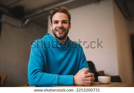 Half length portrait of cheerful male customer smiling at camera during free time in cafe interior, joyful Caucasian hipster guy 20 years old holding caffeine beverage posing in local coffee shop