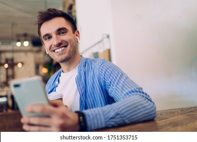 Half length portrait of funny Caucasian man using mobile phone for searching music for listen on entertainment, cheerful male millennial 20 years old in bluetooth headphones enjoying sound records