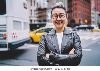Half length portrait of joyful female tourist in optical eyewear and terndy leather jacket posing at urban street, funny Japanese hipster girl with crossed hands smiling at camera during vacations