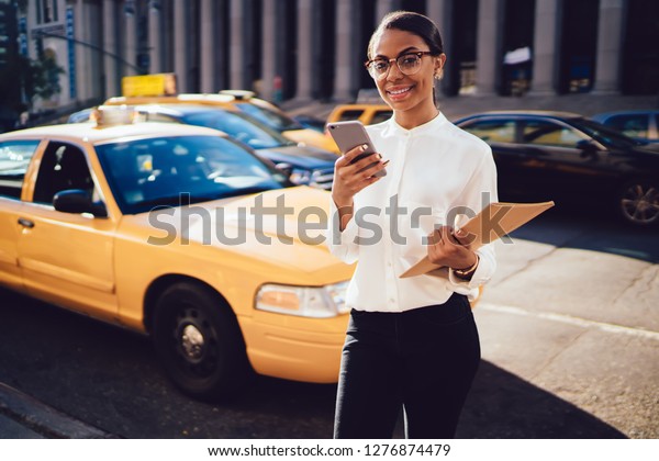Half length portrait of positive female proud ceo\
in spectacles and formal wear holding smartphone gadget and folder\
with documents and looking at camera while waiting yellow cab on\
urban setting