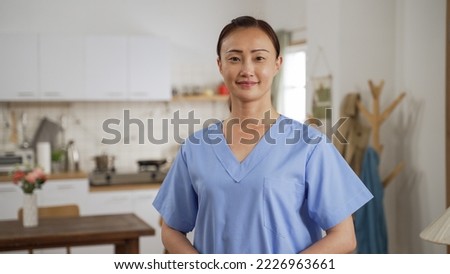 half length portrait of pretty female asian nursing aide wearing uniform looking at camera with smile and folded arms against patient’s house background