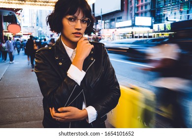 Half Length Portrait Of Stylish Hipster Girl In Cool Eyeglasses Standing Outdoors In Downtown With Time Lapse Of Crowd, Attractive Young Woman Dressed In Leather Jacket Looking At Camera On New York