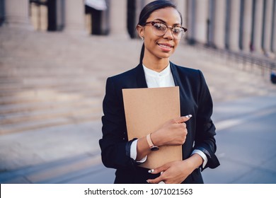 Half length portrait of successful smiling african american female employee dressed in stylish suit holding folder and smiling at camera.Positive dark skinned businesswoman standing in urban setting