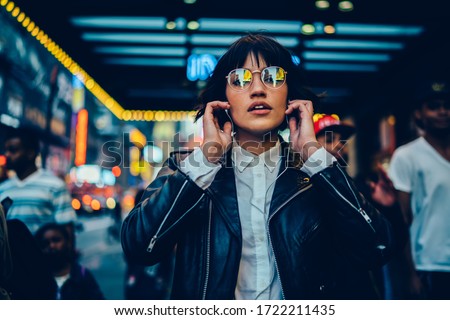Half length portrait of trendy female millennial in electronic spectacles looking at camera during time for listening audio book and walk in metropolitan downtown, generation z in earphones