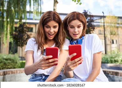 Half length portrait of two brunette twins wearing white t shirts, holding smartphones and looking shocked and surprised. Girls posing with open mouth. People emotions concept - Powered by Shutterstock