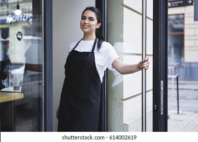 Half length portrait of waitress dressed in black apron with copy space for brand name open store starting work ready to serve visitors, woman entrepreneur barista standing at restaurant entrance