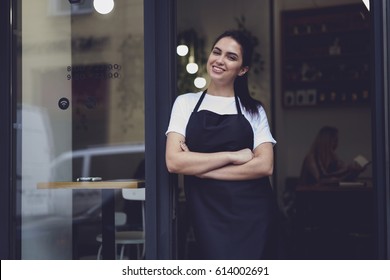 Half Length Portrait Of Young Business Woman Waitress In Black Apron Ready To Attend New Customers In Her Just Opened Coffee Shop.Female Owner Of Restaurant With Smile Standing Near Cafe Door Entrance