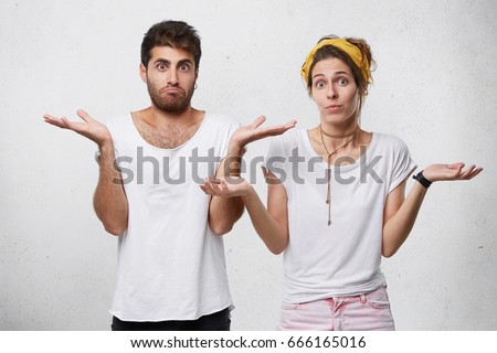 Half length portrait of young couple standing against white background shrugging their shoulders having uncertainty not knowing what to do. Pretty wife and her husband having doubts isolated