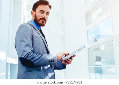 Half Length Portrait Of A Young Serious Man Bookkeeper Holding Touch Pad While Standing In Modern Interior, Handsome Successful Male Office Worker In Suit Using Digital Tablet During Work Break