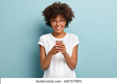 Half length shot of Afro woman holds mobile phone, enjoys nice talk online in social networks, reads funny article in internet, uses cellular, wears white t shirt and spectacles, isolated on blue wall - Shutterstock ID 1407577895