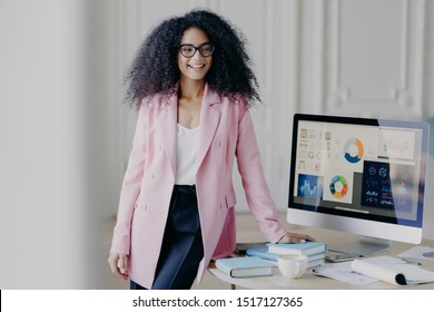 Half length shot of cheerful African American woman with curly hair, dressed in formal elegant outfit, poses near table, computer screen with graphics, prepares for presentation in front of auditorium