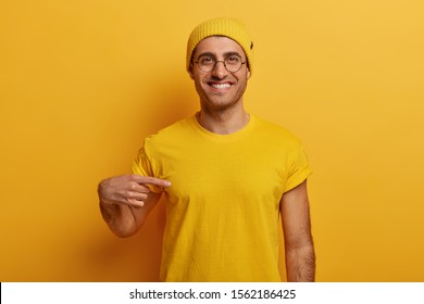 Half Length Shot Of Cheerful Man Points At Mockup Yellow T Shirt, Shows Space For Your Design, Has Glad Expression, Advertises New Outfit, Poses Against Bright Background. Look At This T Shirt