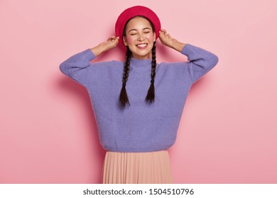 Half length shot of happy Asian woman touches beret, smiles pleasantly, shows white teeth, has two pigtails, wears purple jumper and skirt, expresses pleasant feelings, isolated over pink background