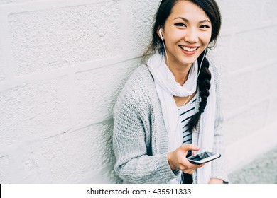 Half length of young beautiful asiatic woman holding a smart phone listening music with earphones, looking in camera, smiling - technology, happiness, music concept