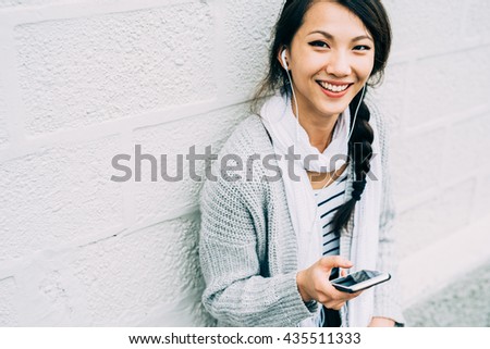 Half length of young beautiful asiatic woman holding a smart phone listening music with earphones, looking in camera, smiling - technology, happiness, music concept