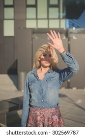 Half Length Of A Young Beautiful Blonde Caucasian Girl Posing In The City Wearing A Jeans Shirt And A Floral Skirt Protecting Her Face From Sun With An Hand 