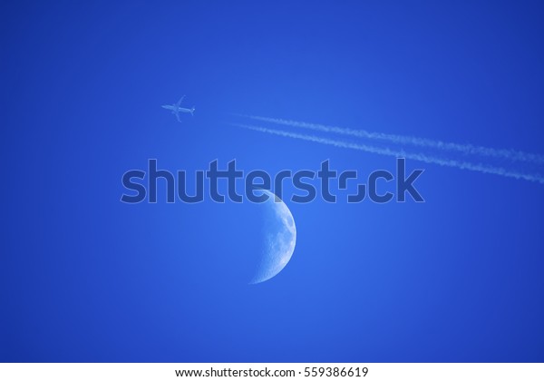 Half\
Moon During Day in Blue Sky. Bright moon orbiting earth in a blue\
clear sky during the daytime with a small plane\
blur