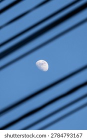 Half moon seen through dark blurred diagonal lines, possibly window blinds or wires. Blue sky background. Image evokes curiosity, mystery, contrast between natural celestial body man-made structure.