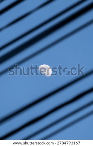 Half moon seen through dark blurred diagonal lines, possibly window blinds or wires. Blue sky background. Image evokes curiosity, mystery, contrast between natural celestial body man-made structure.