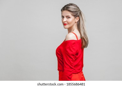 Half Turn Portrait Of Gorgeous Elegant Young Woman With Neat Ponytail Hairstyle In Red Dress Standing, Looking At Camera With Serious Confident Face. Indoor Studio Shot Isolated On Gray Background