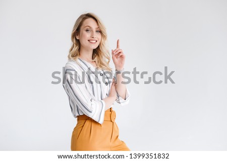 Half turn portrait of young woman wearing casual clothes smiling at camera and pointing finger upward isolated over white background in studio