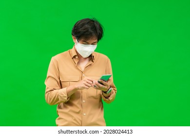 A Half-body Of An Asian Man In A Yellow Shirt Holding His Phone Wearing A Medical Mask Stands Behind A Green Screen.