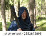 Half-bust portrait of a middle-aged Caucasian man wearing a blue hooded cloak and holding a black wizard staff in one hand, he stands in a wood.
Space for copy.