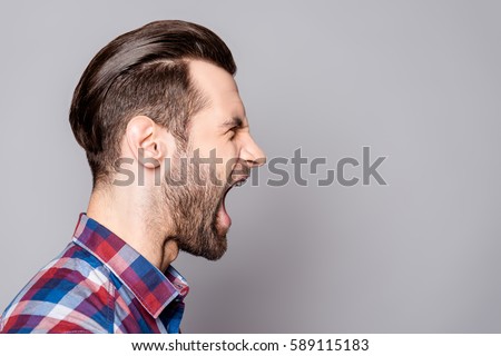 A half-face portrait of a young bearded yelling man with fashionable haircut isolated against gray background.