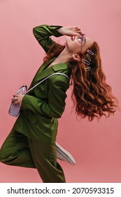 Half-length Happy Caucasian Young Woman In Profile Bends Back With Her Leg Pressed To Knee Indoors. Curly Red Hair Flying Through Air, She Wears Glasses, Suit And Bag On Top.